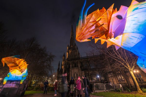 Colorful illuminated inflatable sculptures of animals are displayed at night in front of a tall, gothic-style church, with people walking and gathering around the vibrant artwork in winter clothing.