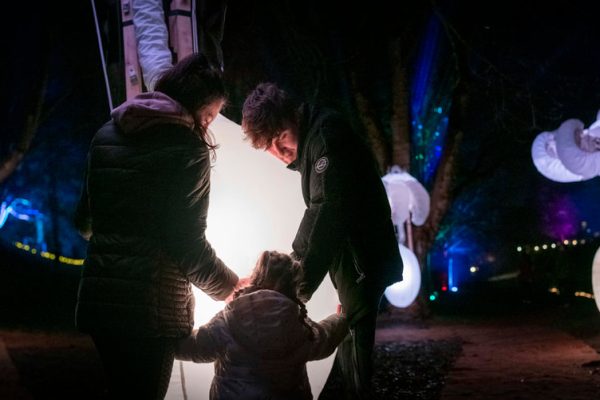 A family with a small child stands near a glowing light installation in a dark, outdoor forest area, surrounded by illuminated, inflatable cloud-shaped decorations hanging from trees.