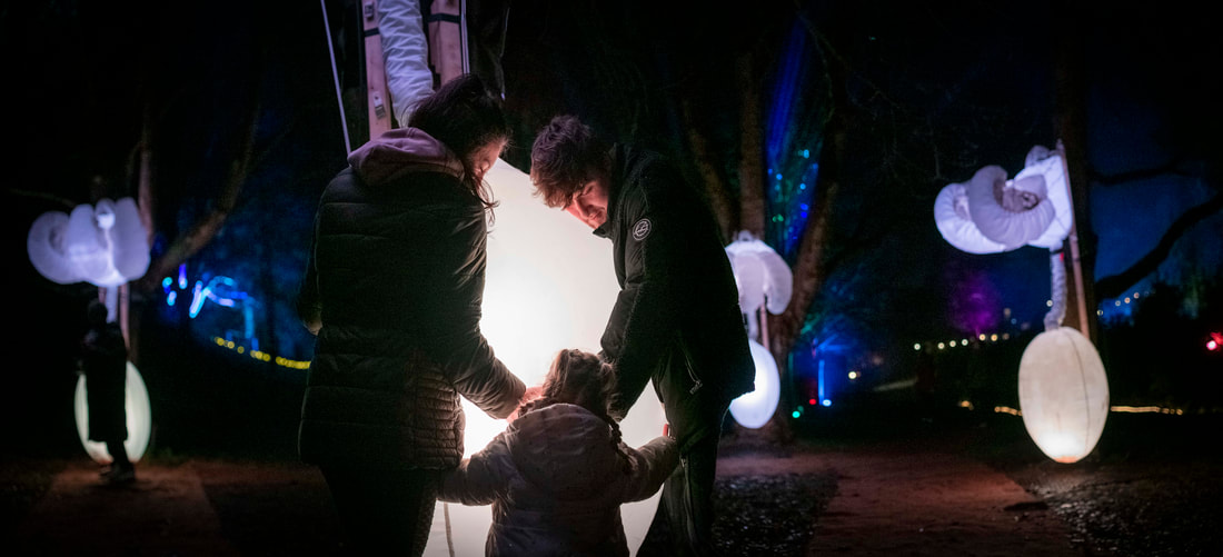A family with a small child stands near a glowing light installation in a dark, outdoor forest area, surrounded by illuminated, inflatable cloud-shaped decorations hanging from trees.