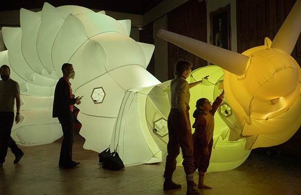 Four people, including a child, interact with a large, illuminated, segmented sculpture resembling a glowing caterpillar or insect inside a dimly lit room. The sculpture emits white and yellow light.